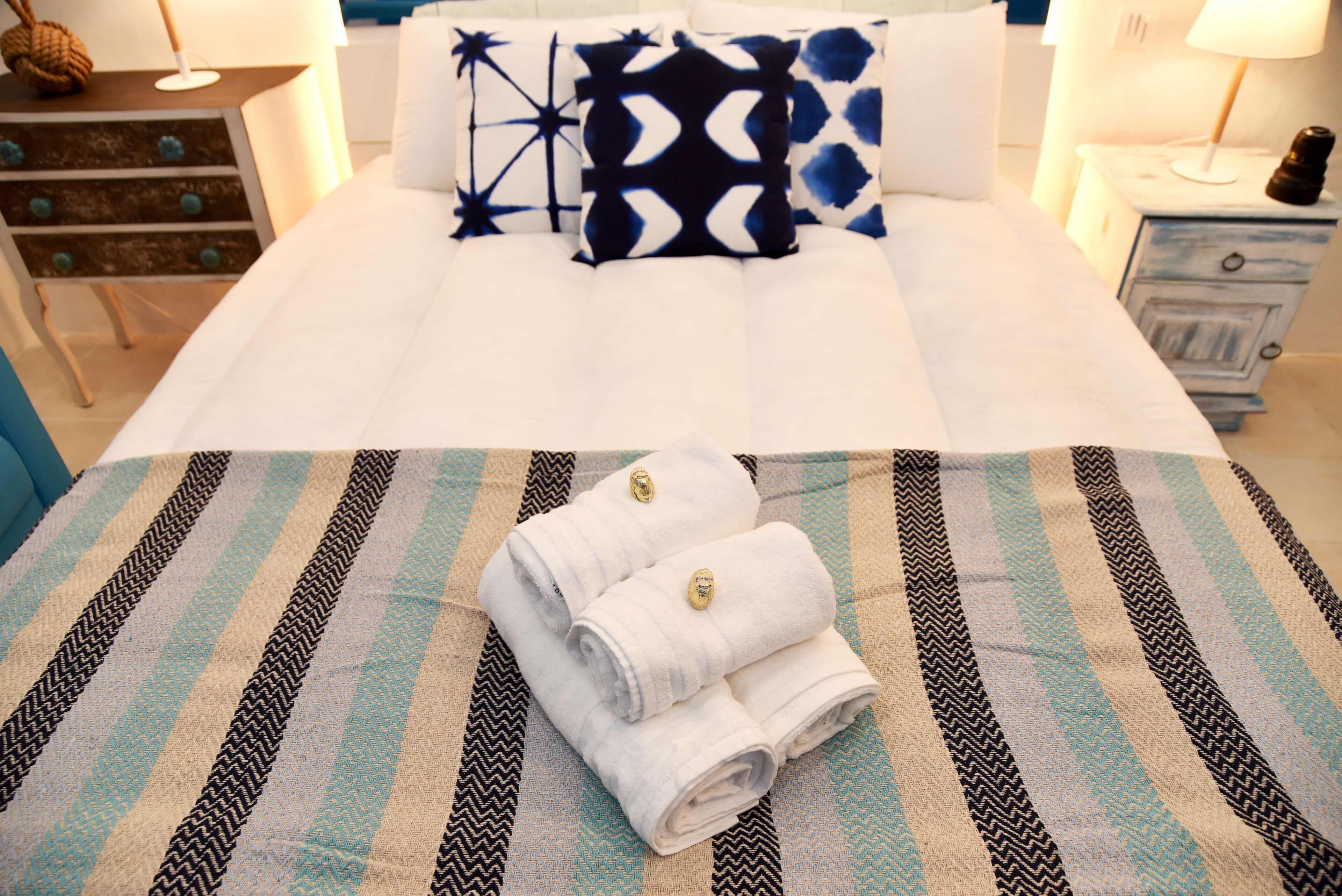 A neatly made bed with white linens and two rolled towels on a striped bedspread.