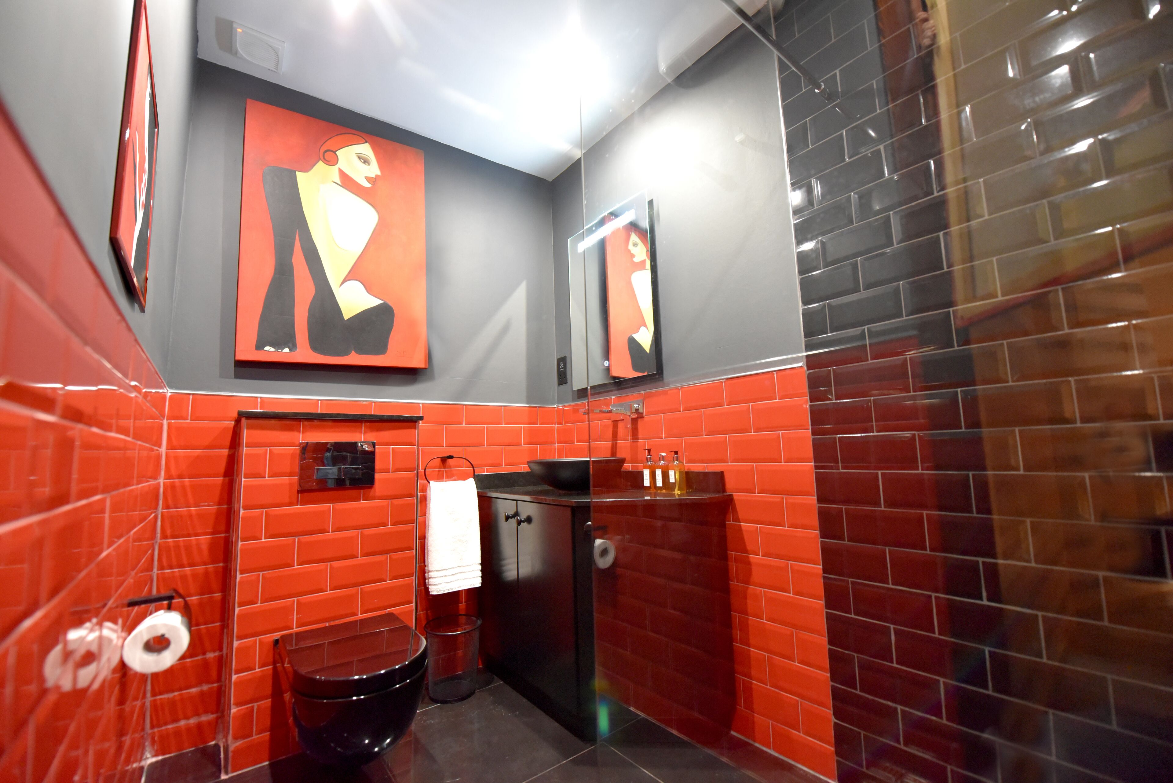 Un baño con paredes cubiertas de azulejos rojos y grises, un inodoro negro, un lavabo con espejo y una toalla colgada en la pared.
