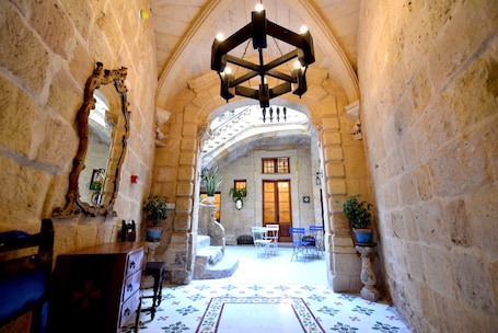 A stone-walled corridor with a wooden bench, a mirror, and a chandelier. There are potted plants and a staircase leading to a room with a wooden door.