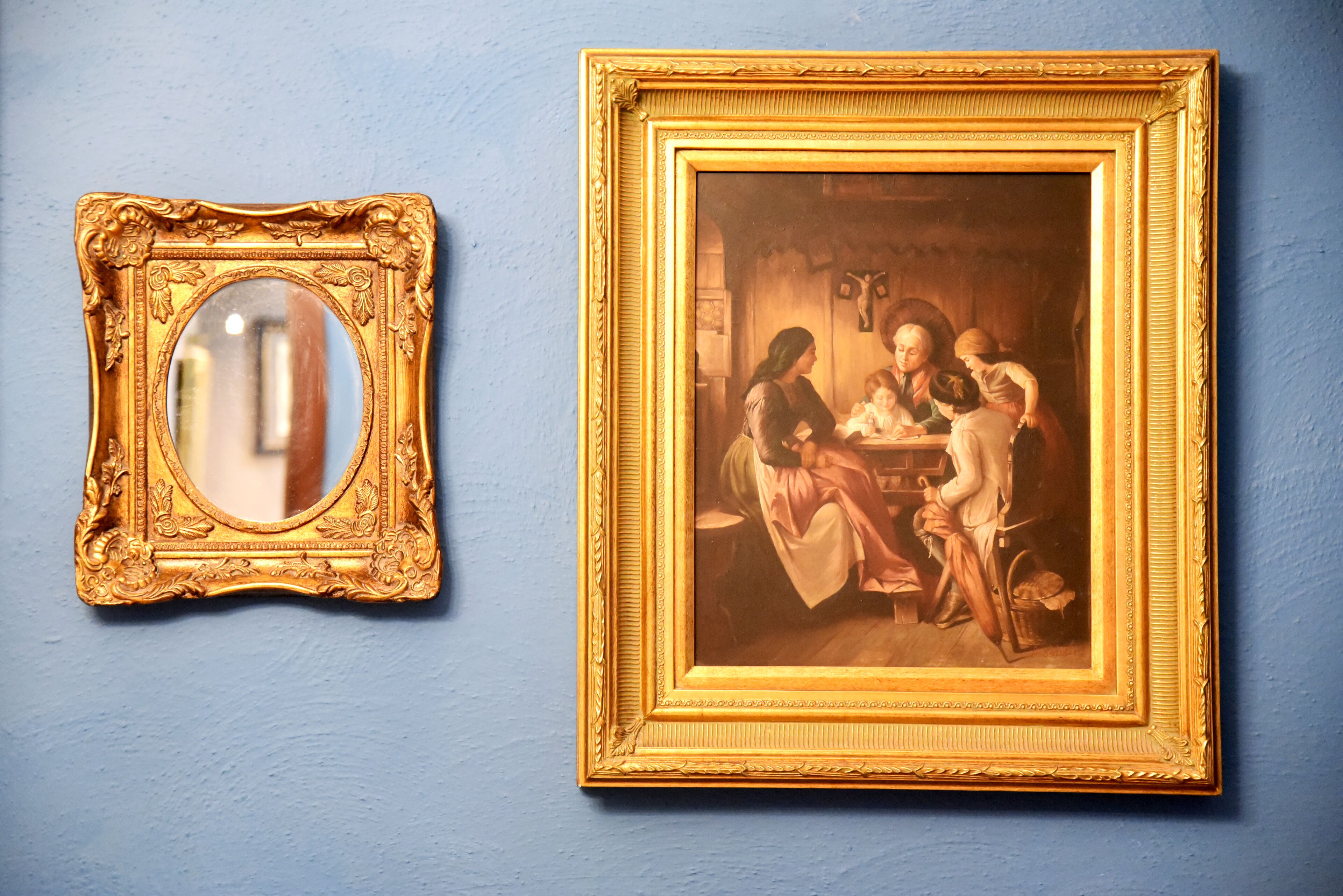 A room with a blue wall, a large framed painting, and a mirror with an ornate gold frame.