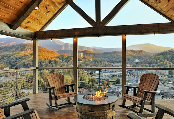 Une terrasse en bois avec un foyer en pierre et deux fauteuils Adirondack, offrant une vue sur un paysage montagneux.