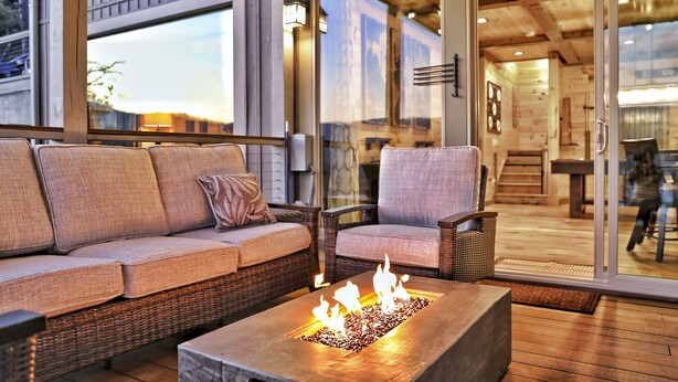 Un área de estar al aire libre con una chimenea, mobiliario de mimbre y un ventanal que da a un espacio interior.