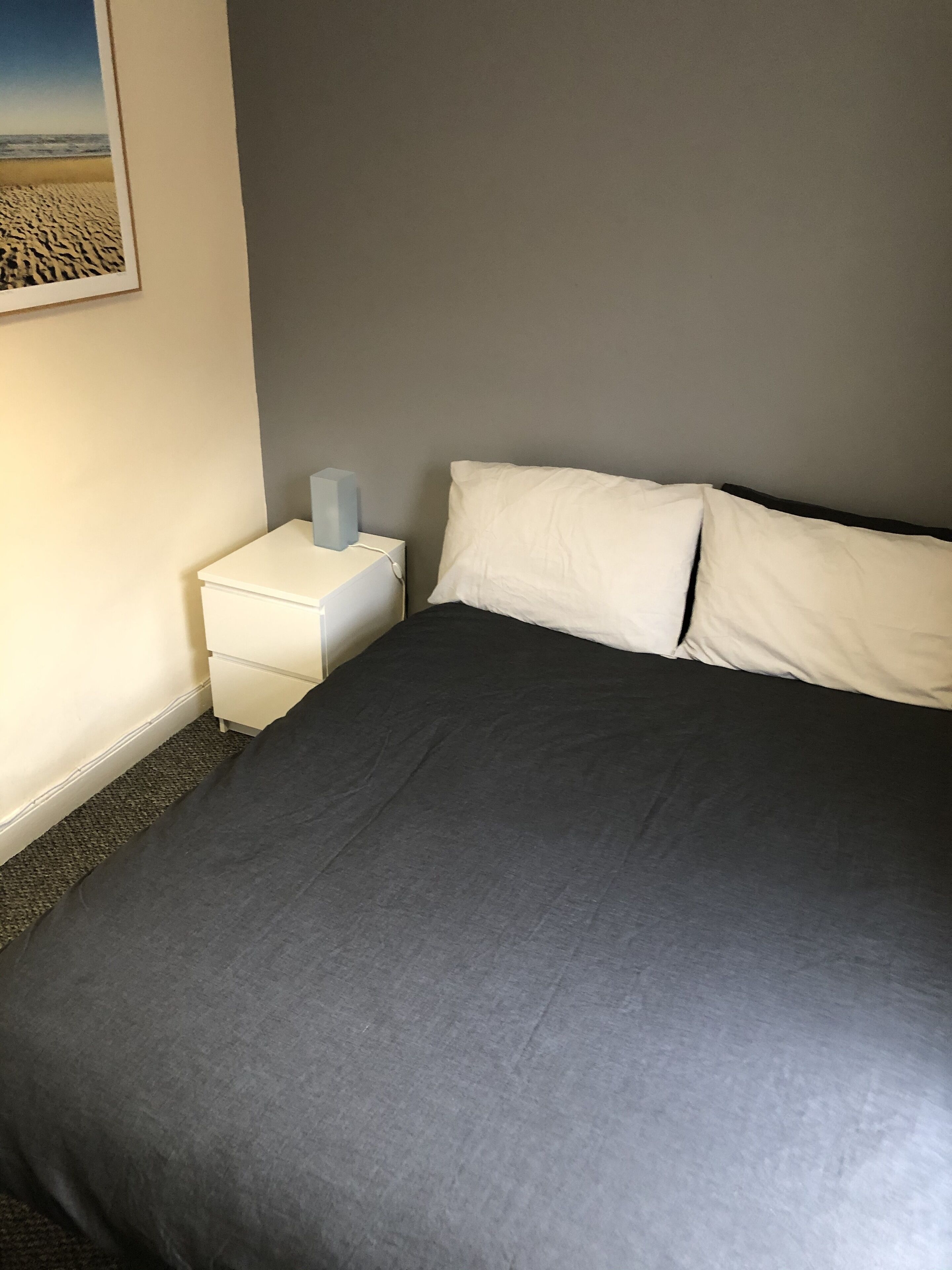 Un lit bien fait, recouvert d’une couvre-lit gris foncé et agrémenté d’oreillers blancs, une table de chevet blanche avec une lampe, et un tableau encadré représentant une plage, accroché au mur.