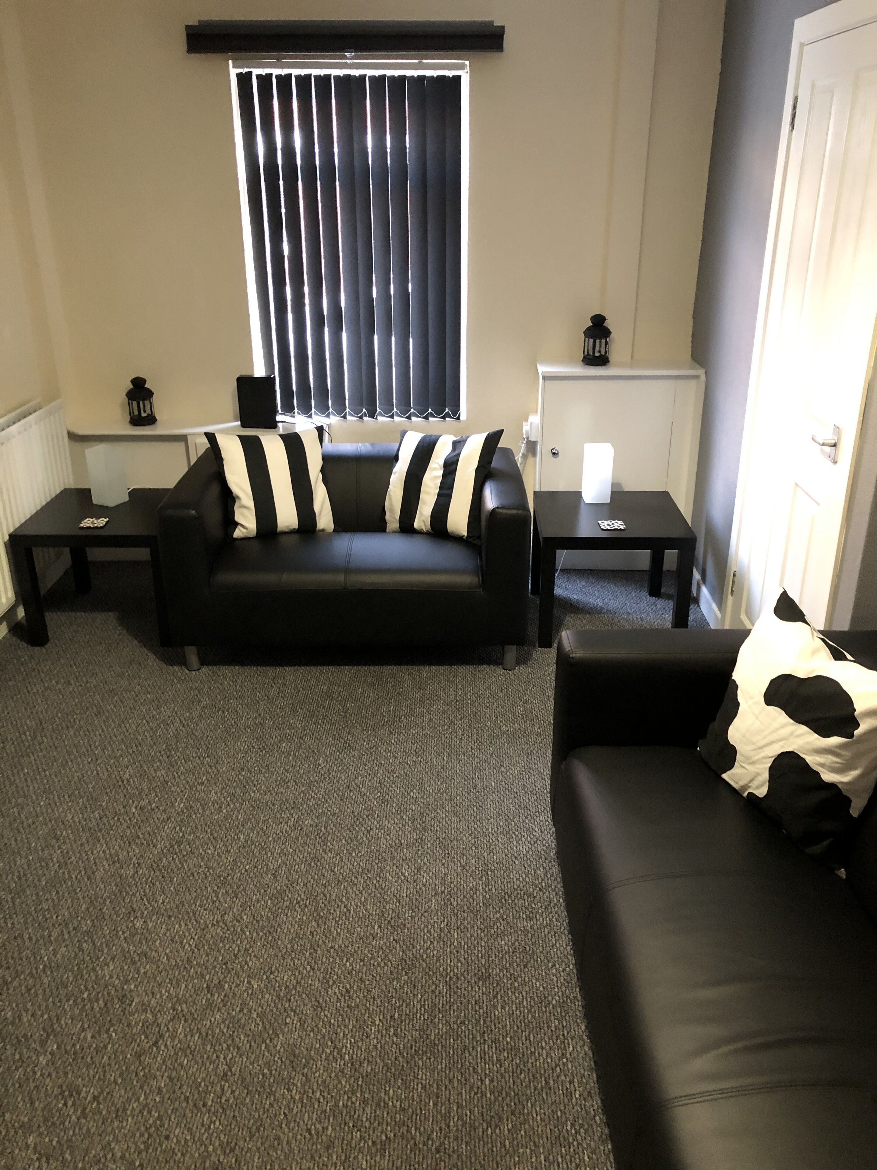 A living room with a black leather sofa, a black and white striped pillow, a black and white cow print pillow, and a black leather couch. There are two black side tables with a white lamp on each. A window with vertical blinds is visible in the background.