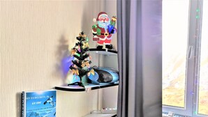 Eine Ecke eines Zimmers mit einem kleinen Weihnachtsbaum, einer Weihnachtselfigur, einem Buch mit dem Titel 'Guide' und einem Fenster mit Vorhängen.