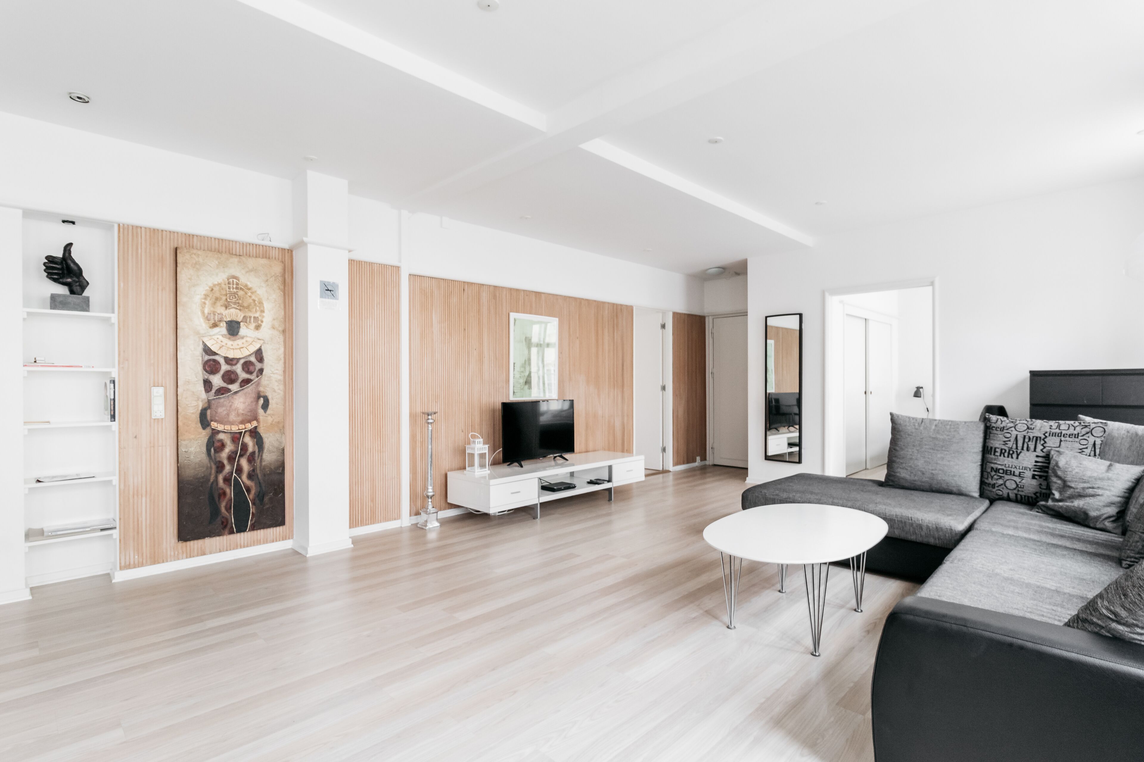 A modern living room with a wooden floor, a white sofa, a white coffee table, a TV on a wooden cabinet, and a large abstract painting on the wall.
