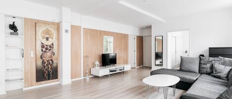 A modern living room with a wooden floor, a white sofa, a white coffee table, a TV on a wooden cabinet, and a large abstract painting on the wall.