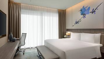 A hotel room with a large bed, a desk with a chair, a bench, a TV, and a painting on the wall.
