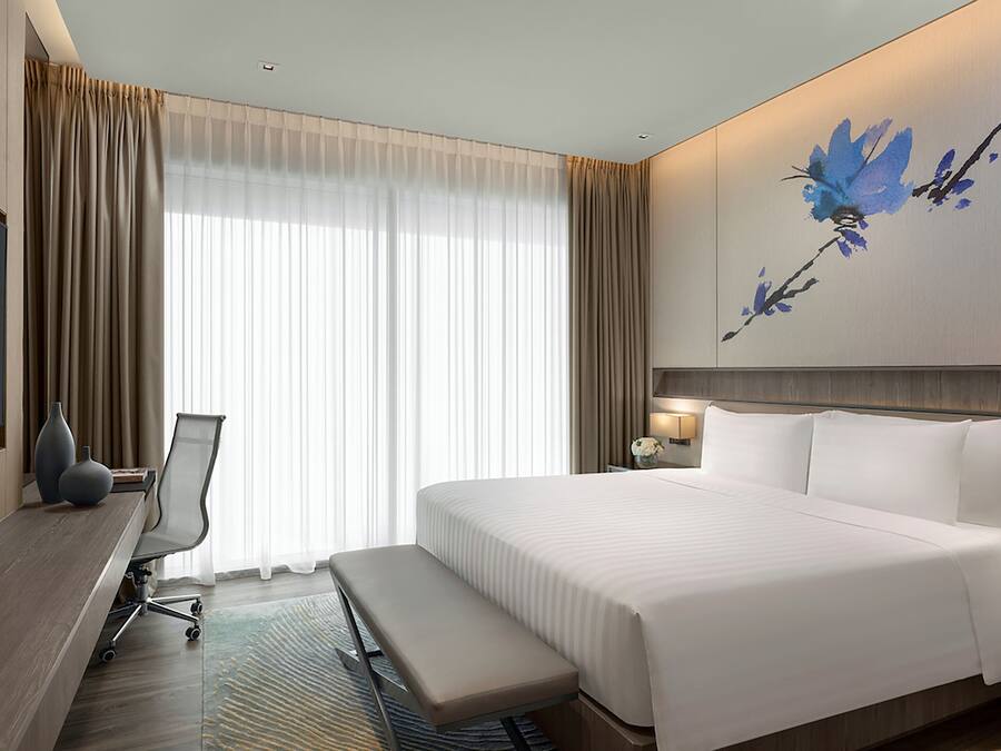A hotel room with a large bed, a desk with a chair, a bench, a TV, and a painting on the wall.