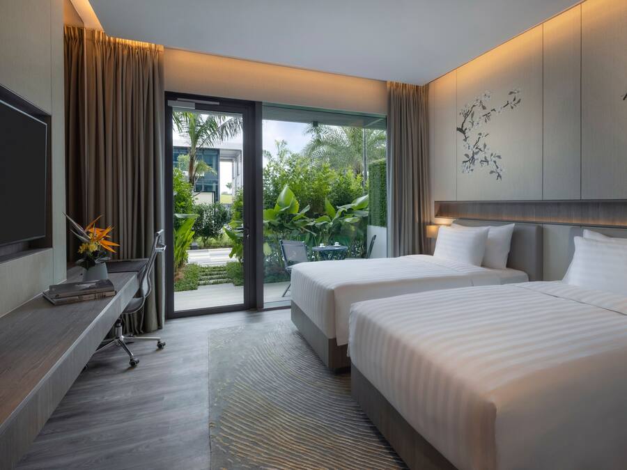 Habitación de hotel moderna con dos camas, un escritorio y un gran ventanal corredizo que da a un balcón con vegetación.