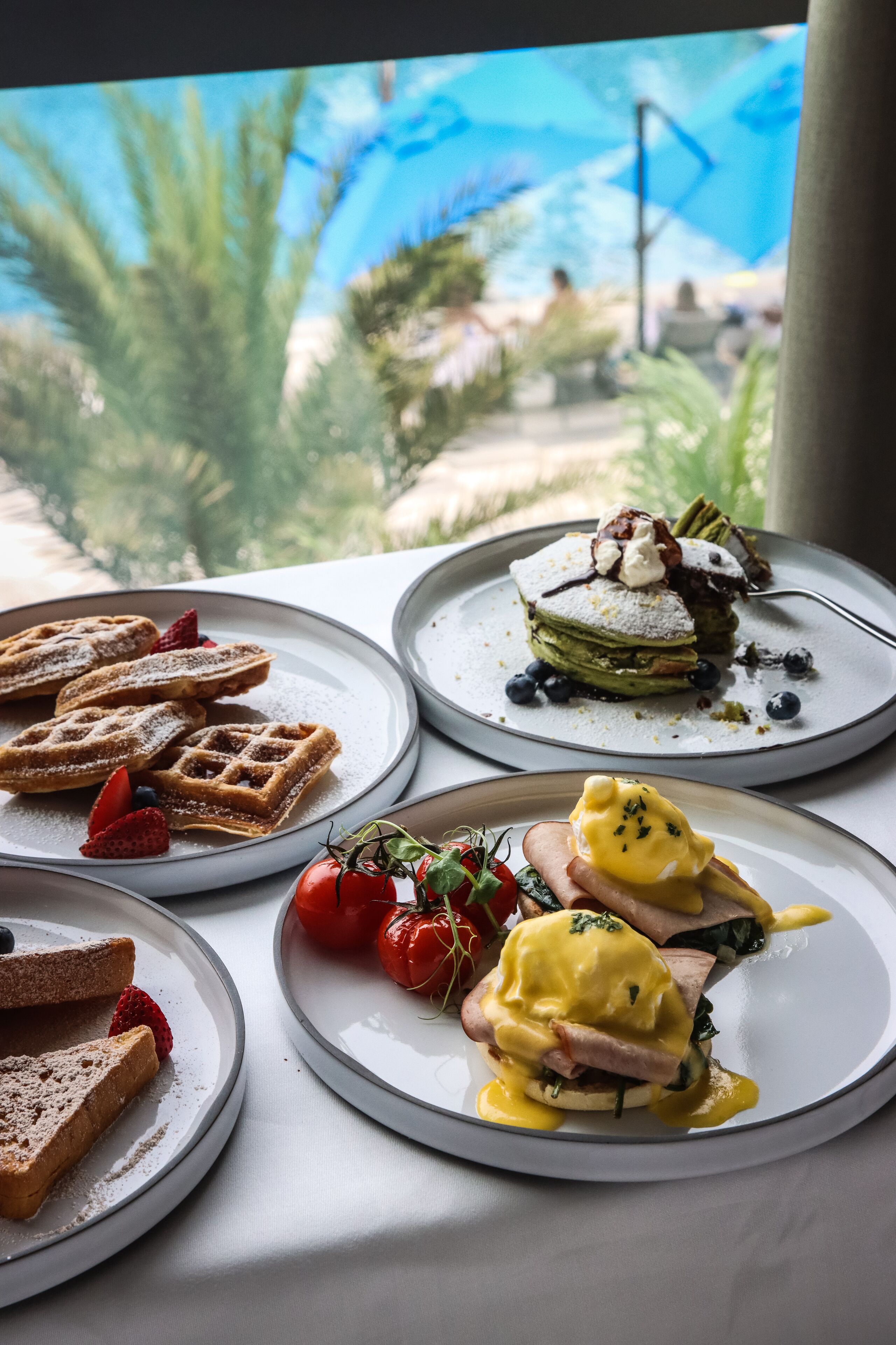 A table with various breakfast dishes, including waffles, a layered dessert, and eggs Benedict, with a view of greenery and a building in the background.