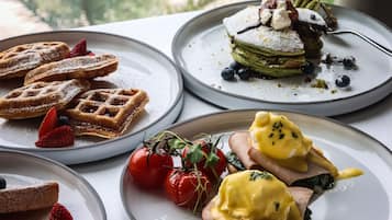 A table with various breakfast dishes, including waffles, a layered dessert, and eggs Benedict, with a view of greenery and a building in the background.