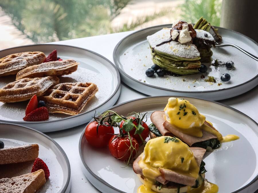 A table with various breakfast dishes, including waffles, a layered dessert, and eggs Benedict, with a view of greenery and a building in the background.