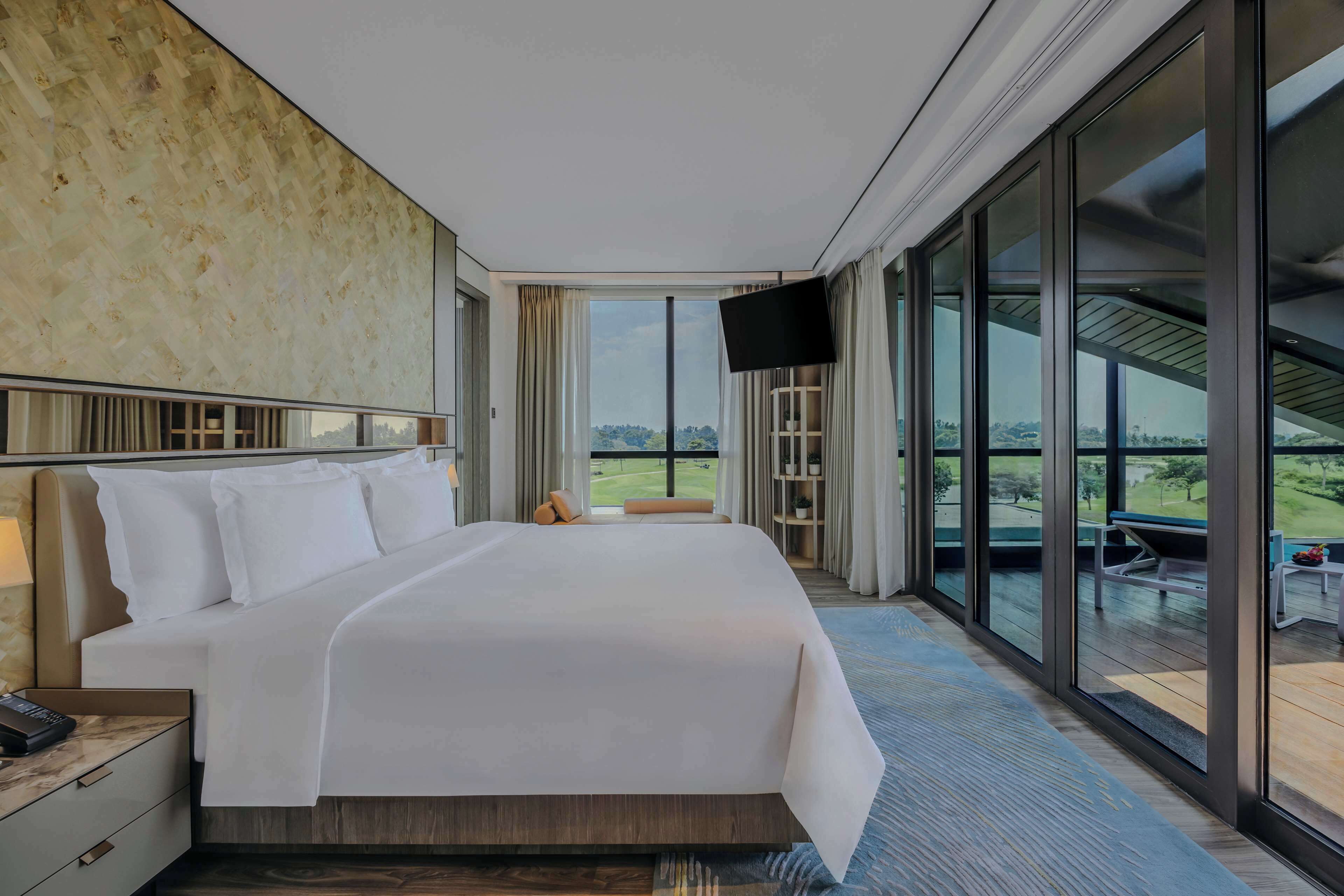 A modern hotel room with a large bed, a flat-screen TV, and a balcony with a view of the landscape.