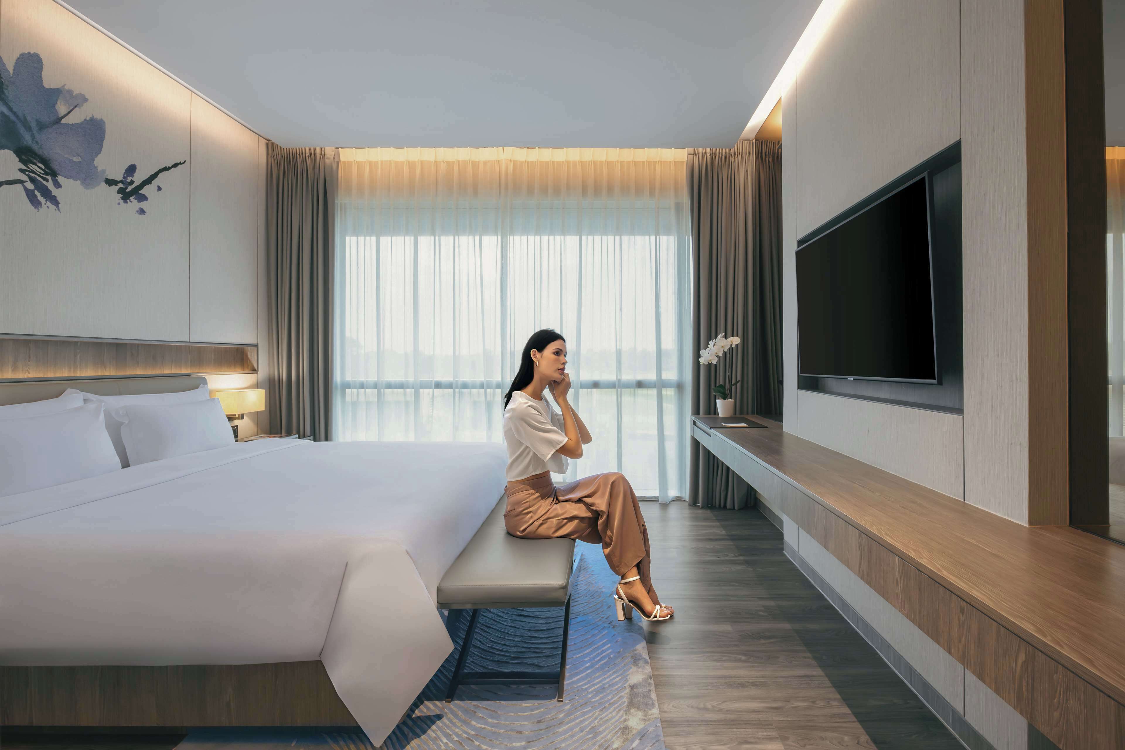 A modern hotel room with a large bed, a flat-screen TV mounted on the wall, and a seating area with a view of the city.