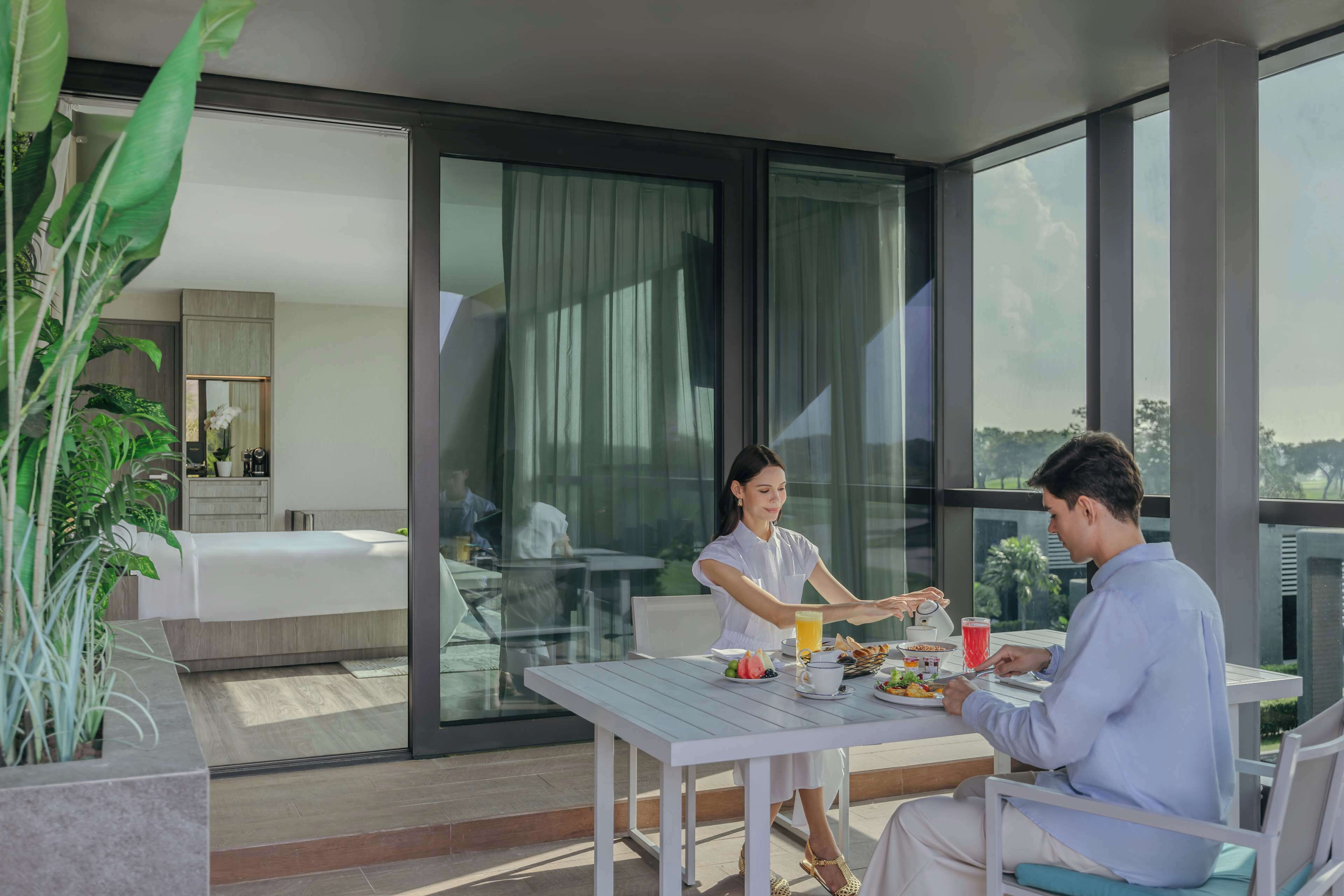 A modern hotel room with a balcony, a dining table with breakfast, and a large window with a view of the outdoors.