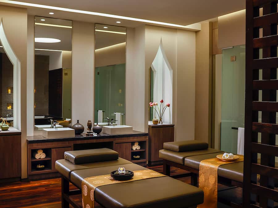 A modern spa room with a large mirror, two massage tables, a sink, and a wooden partition.