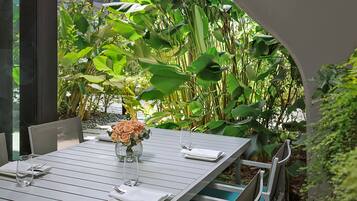 A dining table set with white plates, glasses, and a vase of flowers, surrounded by chairs and greenery.