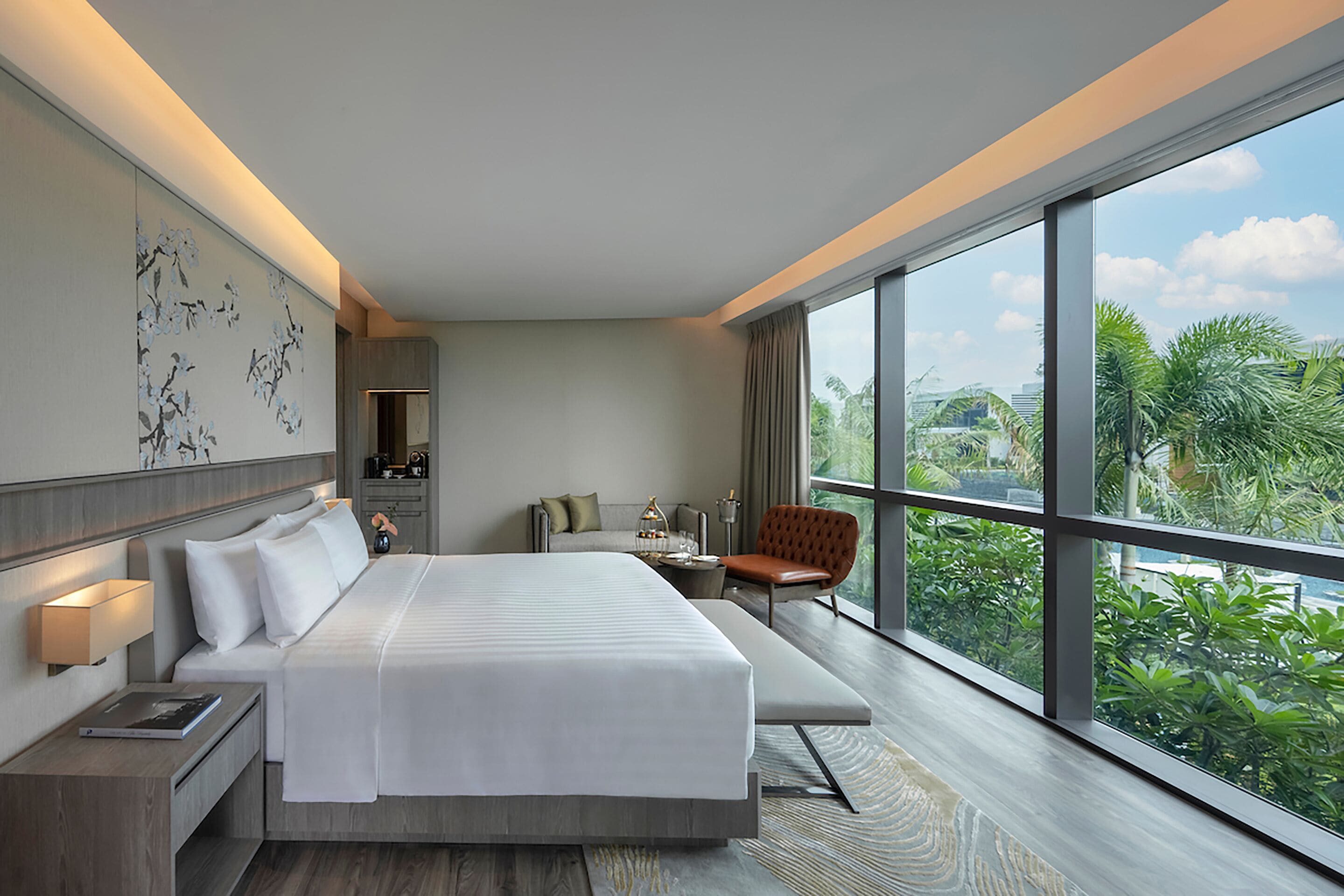 Quarto de hotel moderno com uma cama grande, mesas de cabeceira, um espaço de estar e vista para o verde através de grandes janelas.