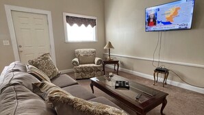 Una sala de estar con un sofá color beige, un sillón estampado, una mesa de centro de madera y un televisor en la pared mostrando un mapa.