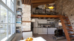 A kitchen with white cabinets, a wooden ceiling, and a stone wall. There is a window on the left, a table with a bowl of fruit, and a ladder leading to a loft area with bookshelves. - Live in the Shadow of the Acropolis (Athens)