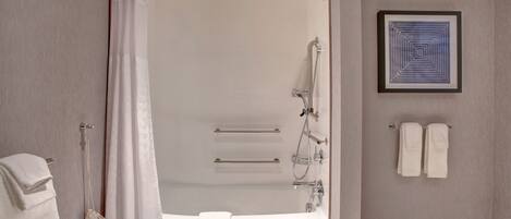 A modern bathroom with a bathtub, shower, and handrails. There are towels and a framed artwork on the wall.
