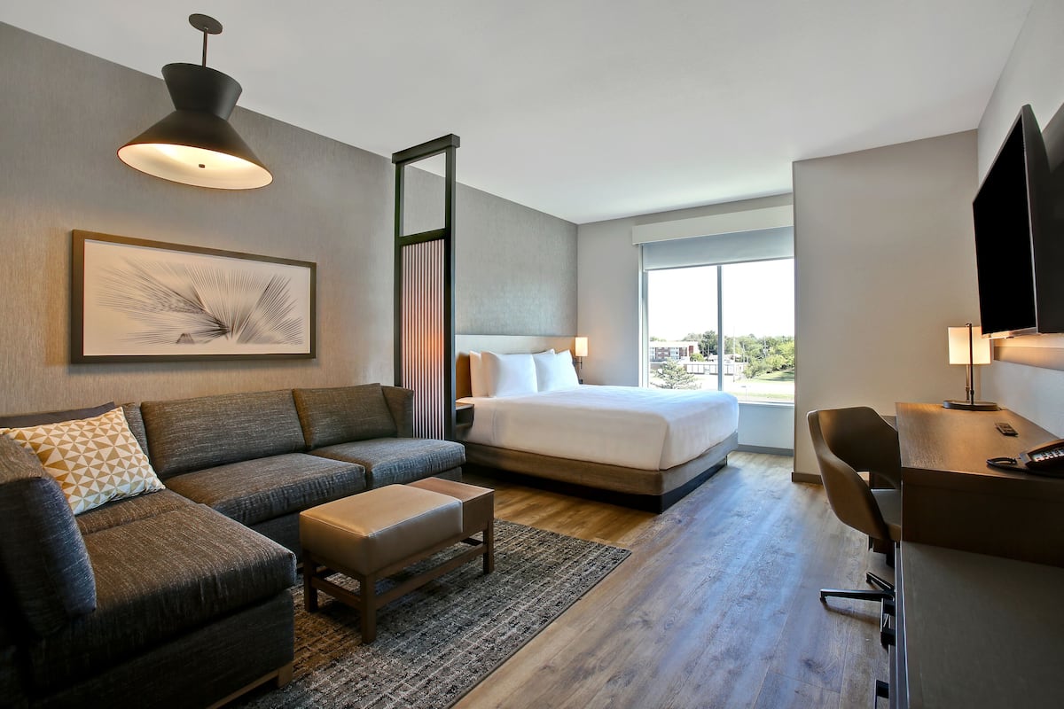 A modern hotel room with a large bed, a sofa, a desk, and a view of the outdoors.