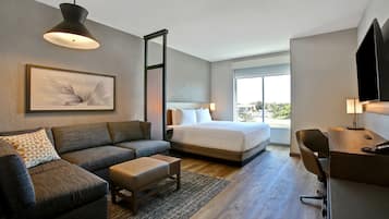 A modern hotel room with a large bed, a sofa, a desk, and a view of the outdoors.
