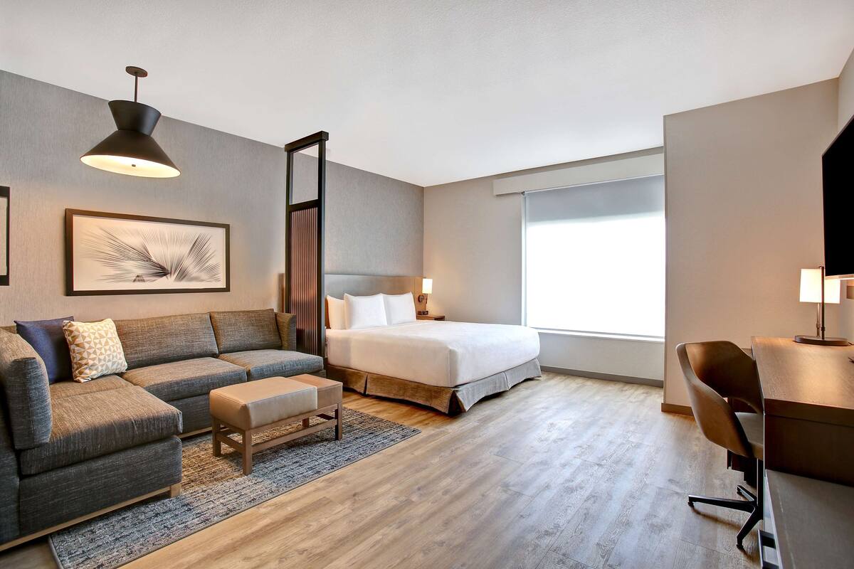 A modern hotel room with a bed, a sofa, a desk, and a large window.