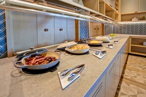 Disposição de buffet com diversos pratos, incluindo um grande recipiente com bacon, tigelas de comida e utensílios de servir.