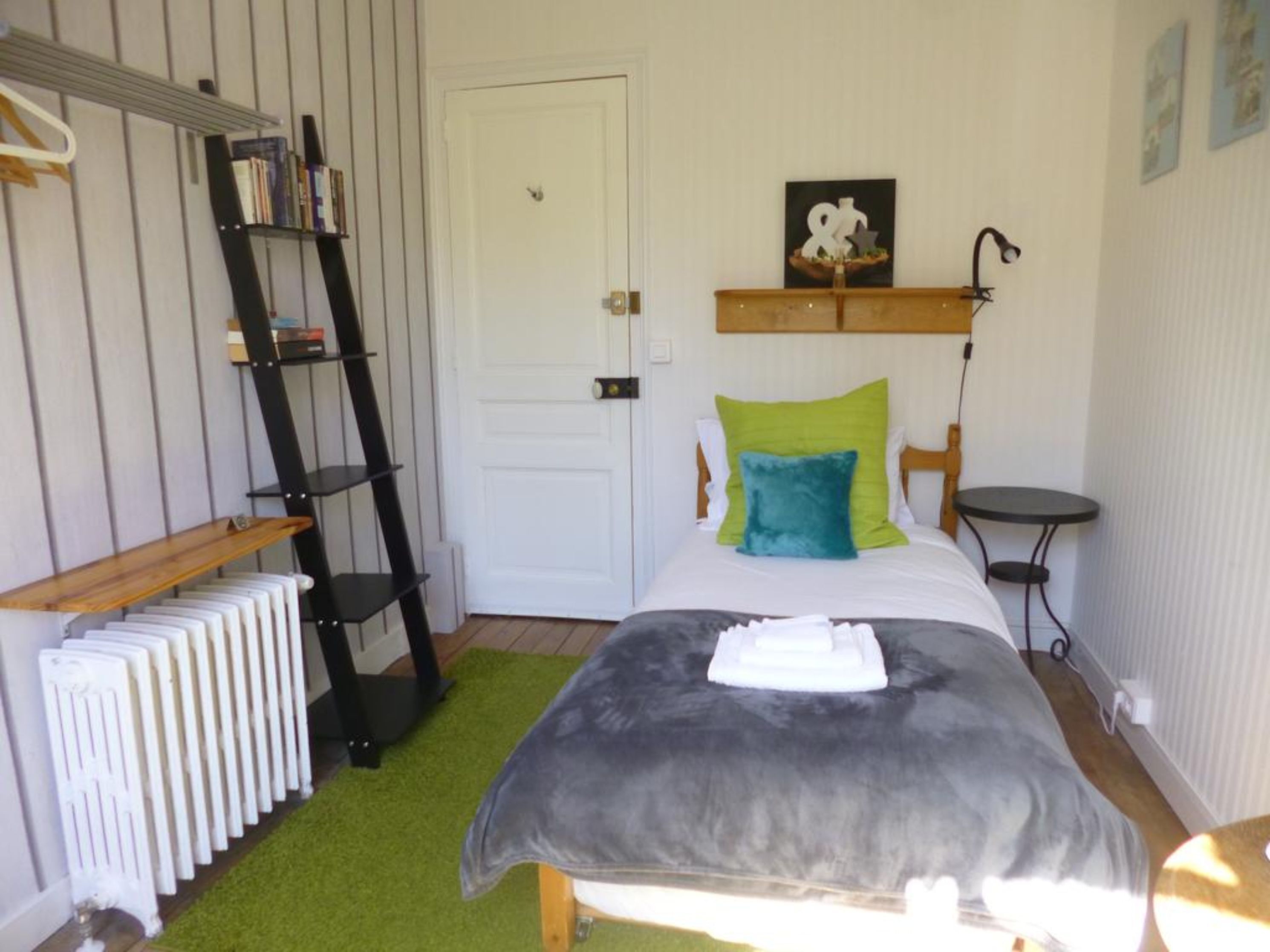 Une chambre à coucher avec un lit, un radiateur, une étagère remplie de livres, une petite table et un tableau accroché au mur.