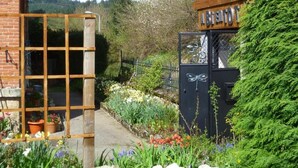 Une entrée de jardin agrémentée d’un portail en bois, d’un portail en métal noir avec une enseigne, ainsi que de diverses fleurs et plantes.