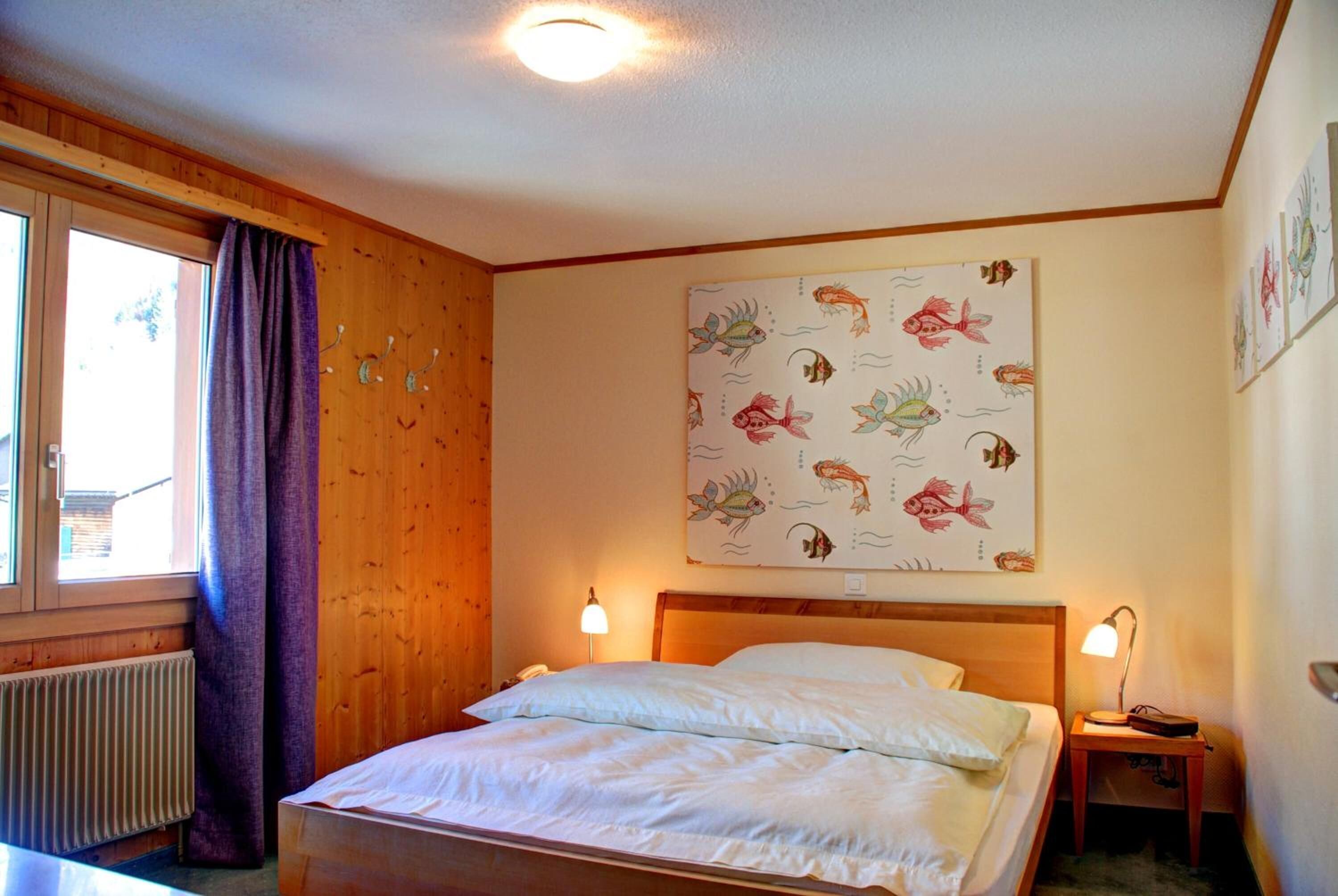 Un dormitorio con cabecera de madera, cama con ropa de cama blanca, ventana con cortinas moradas y pared con estampado de peces.