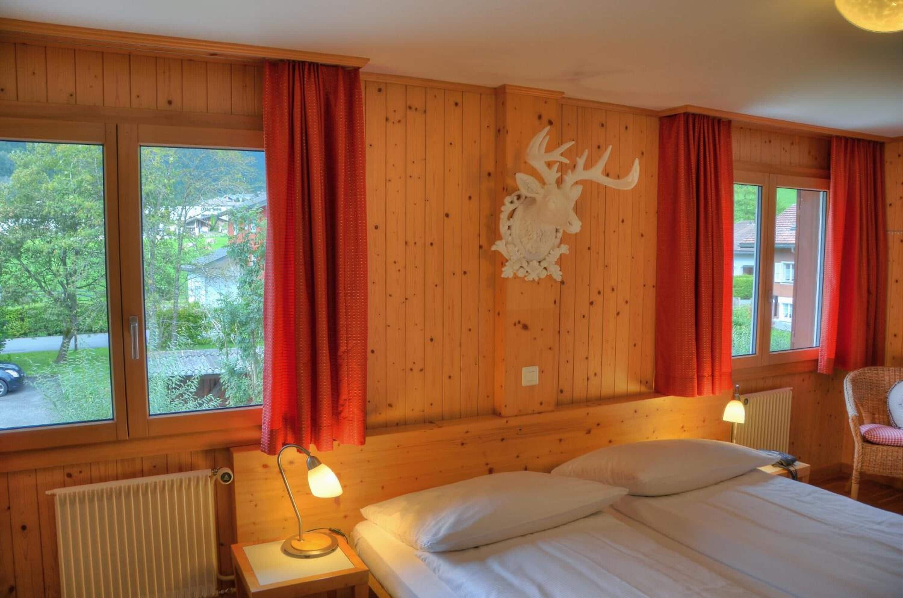 A wooden cabin room with a bed, two bedside lamps, a red curtain, a mounted deer head, and a window with a view of greenery and a building.