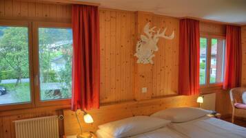 A wooden cabin room with a bed, two bedside lamps, a red curtain, a mounted deer head, and a window with a view of greenery and a building.