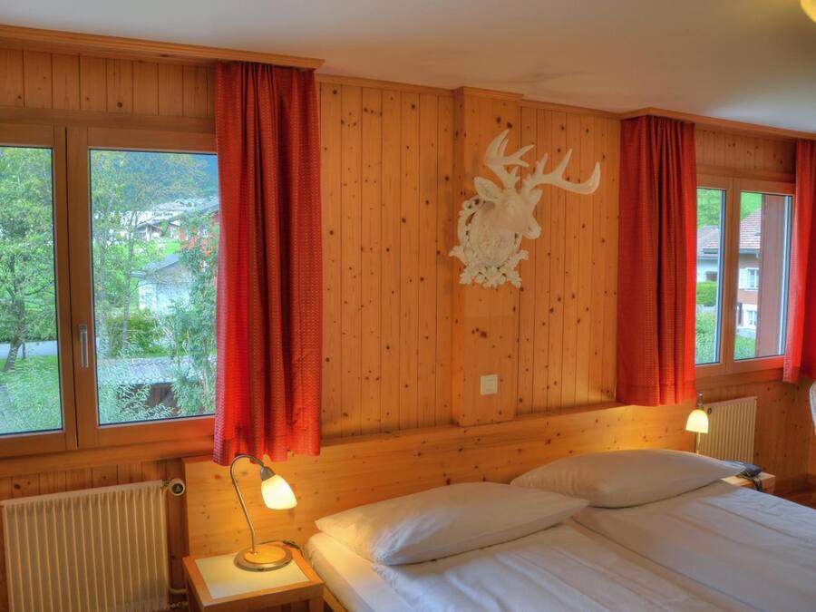 A wooden cabin room with a bed, two bedside lamps, a red curtain, a mounted deer head, and a window with a view of greenery and a building.