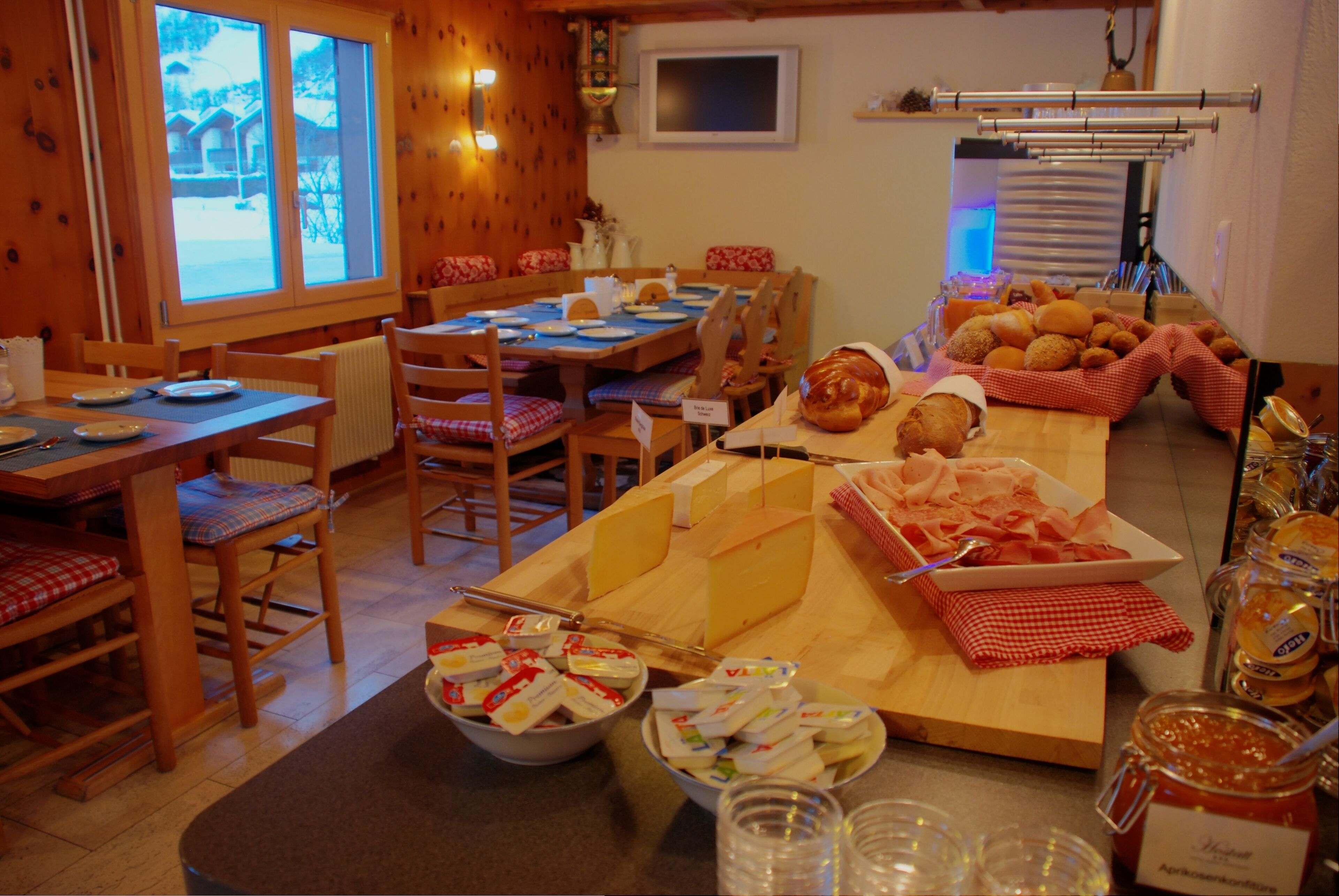 A cozy dining area with wooden furniture, a buffet table with cheese and bread, and a view of the outdoors through large windows.