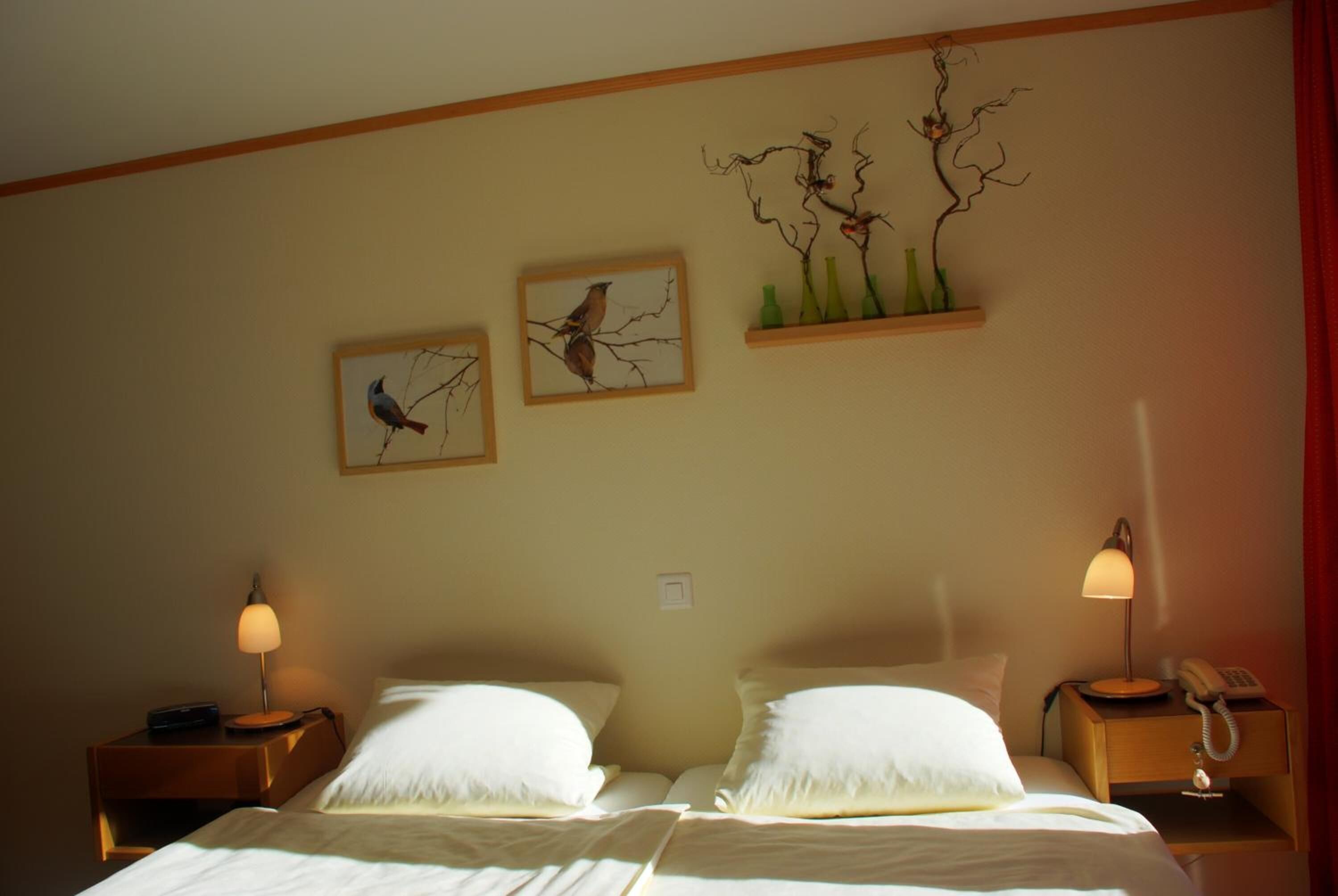 Habitación de hotel con dos camas, una mesita de noche con lámpara y un cuadro con aves.