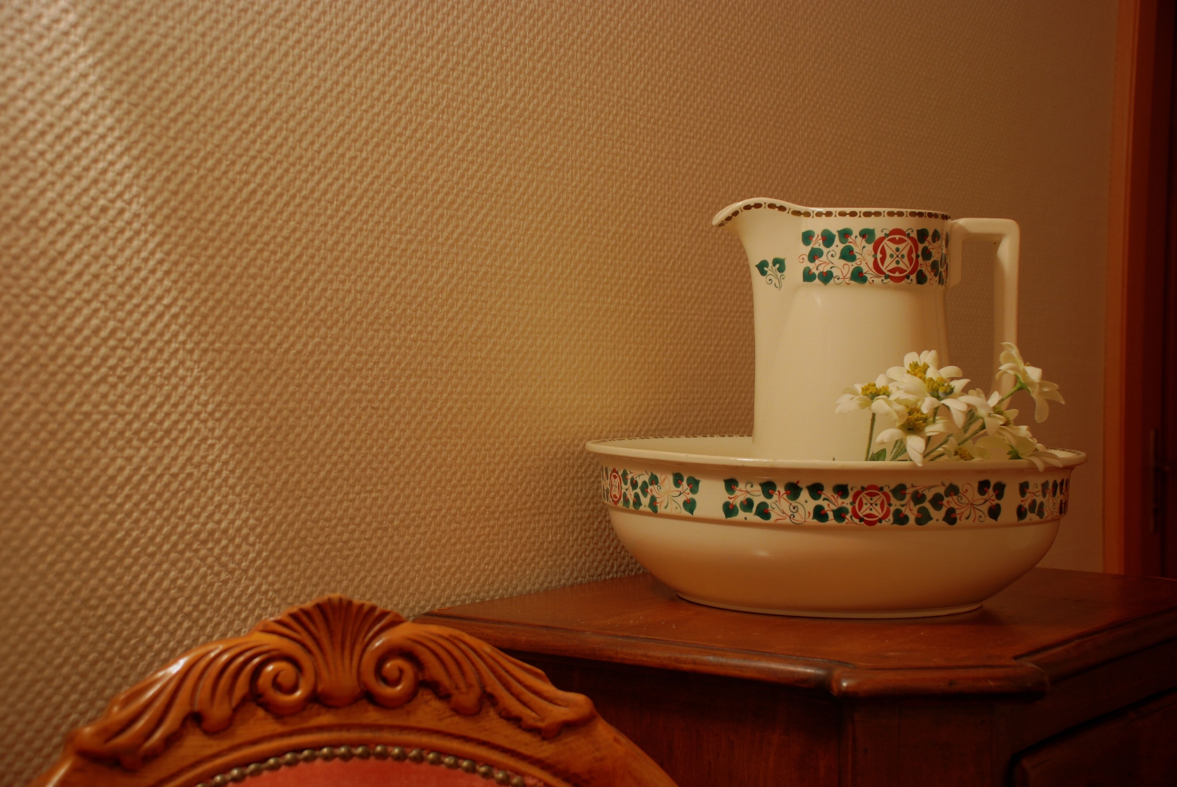 A white pitcher with a floral pattern and a matching bowl on a wooden table against a textured wall.