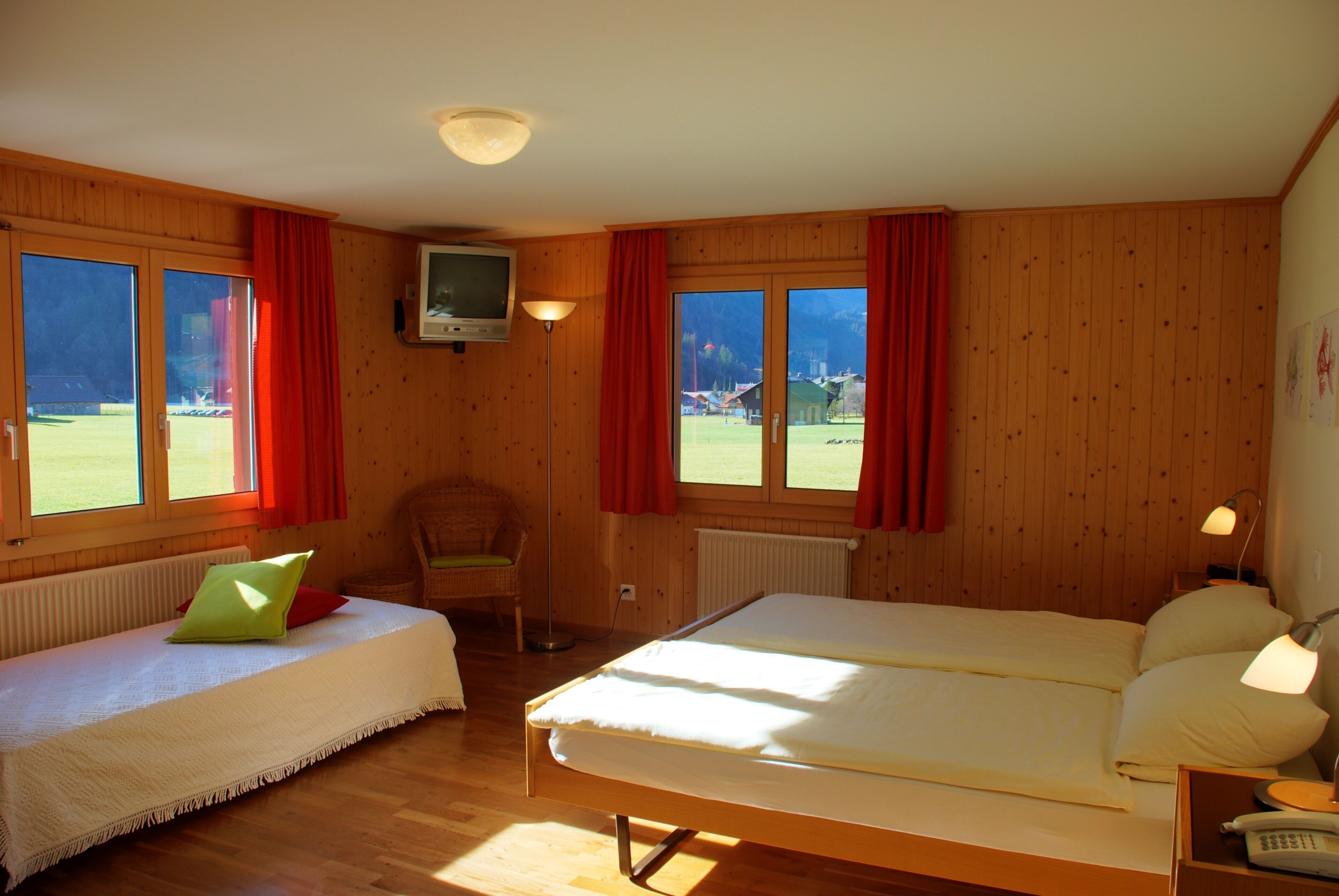Una habitación con cama, sillón, televisor y ventanas amplias con cortinas.