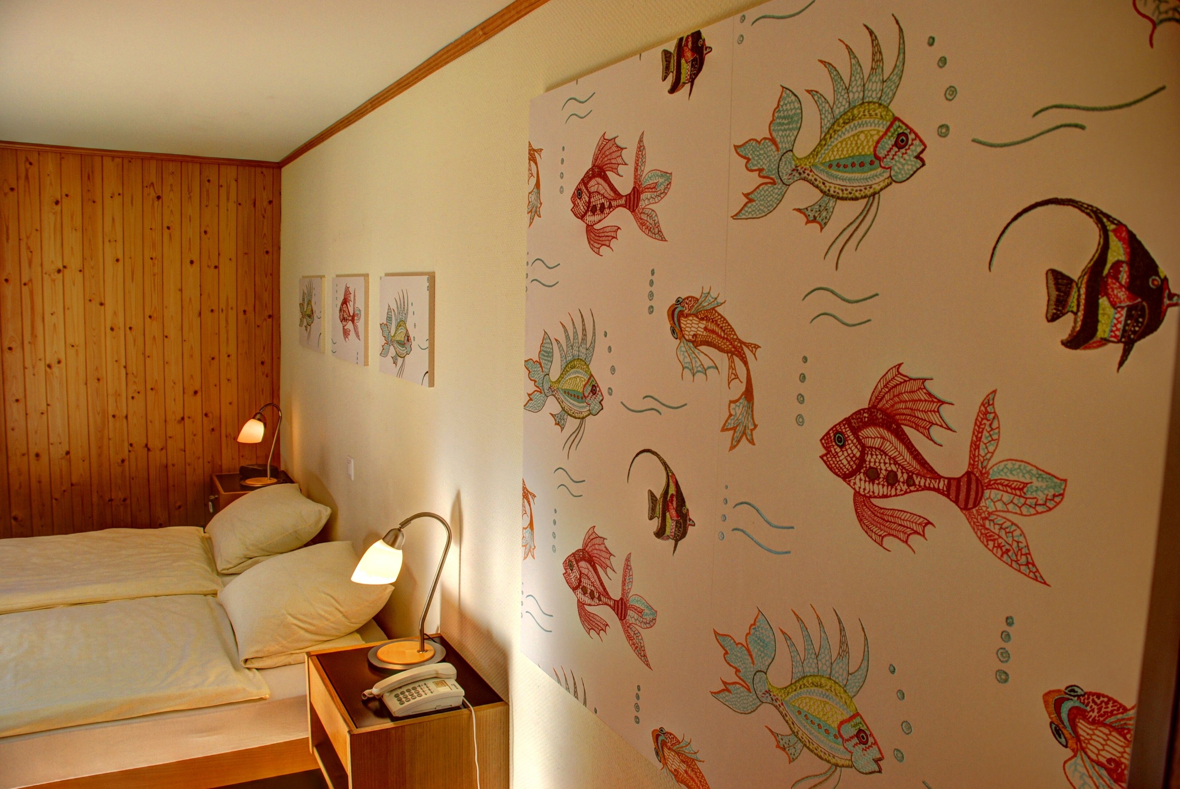 A bedroom with a wooden paneled wall, a bed with two pillows, a bedside table with a lamp and a telephone, and a wall with a fish pattern.