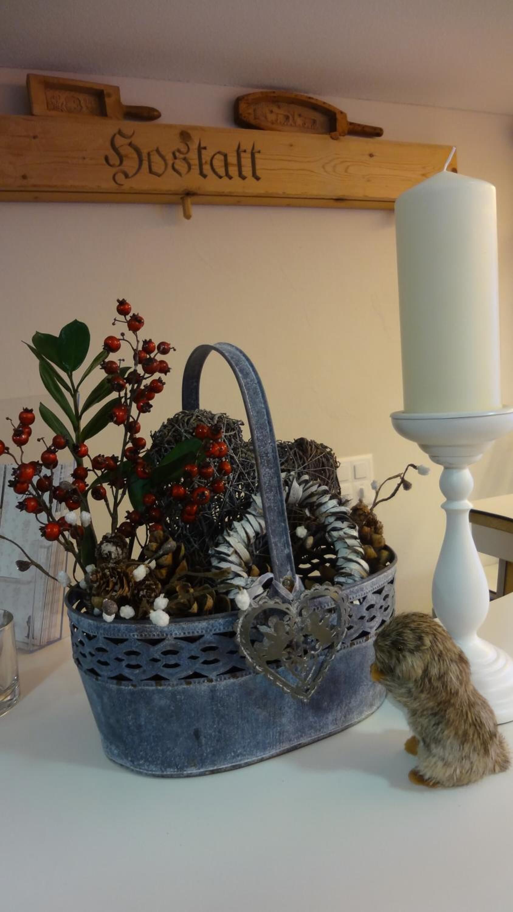 A decorative basket with red berries and pine cones, a white candle on a stand, and a small animal by the basket.