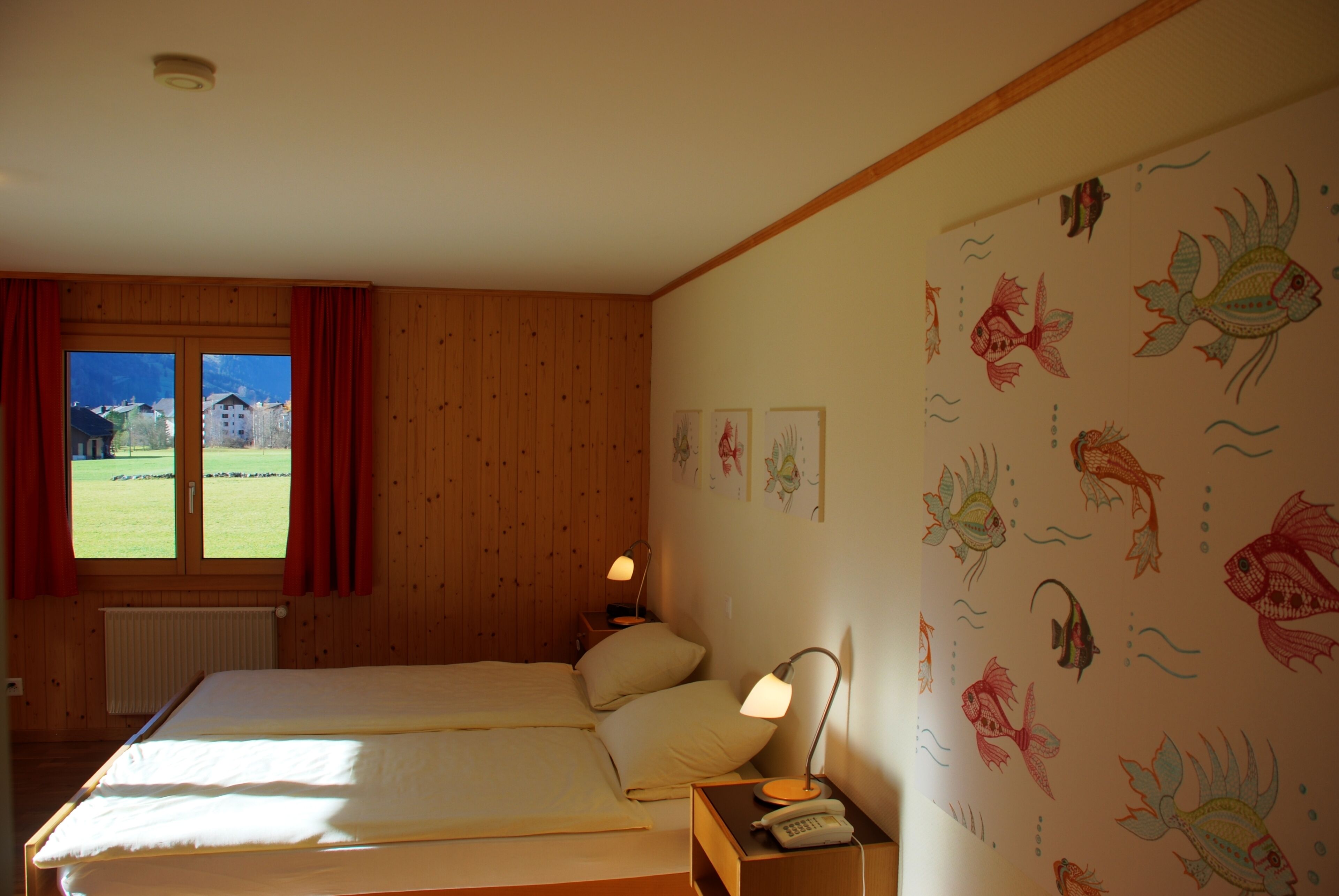 Una habitación con dos camas, un ventanal con cortinas rojas, una pared decorada con ilustraciones de peces y una mesita de noche con una lámpara y un teléfono.