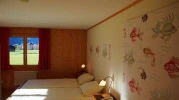 Una habitación con dos camas, un ventanal con cortinas rojas, una pared decorada con ilustraciones de peces y una mesita de noche con una lámpara y un teléfono.