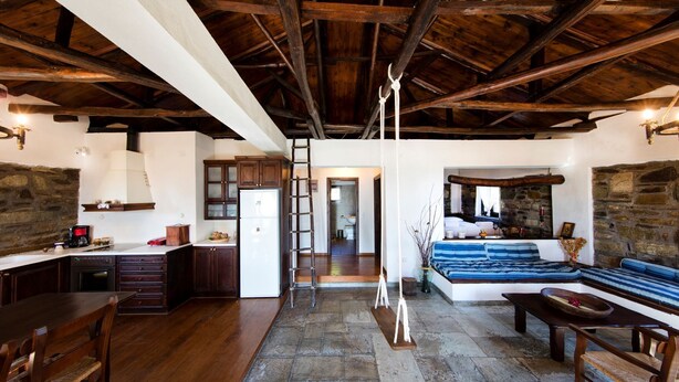 A rustic interior with exposed wooden beams, a stone wall, a kitchen with dark wooden cabinets, a stone floor, and a cozy seating area with a patterned sofa. - Beautiful Tranquil Stone Cottage (Zagora-Mouresi)