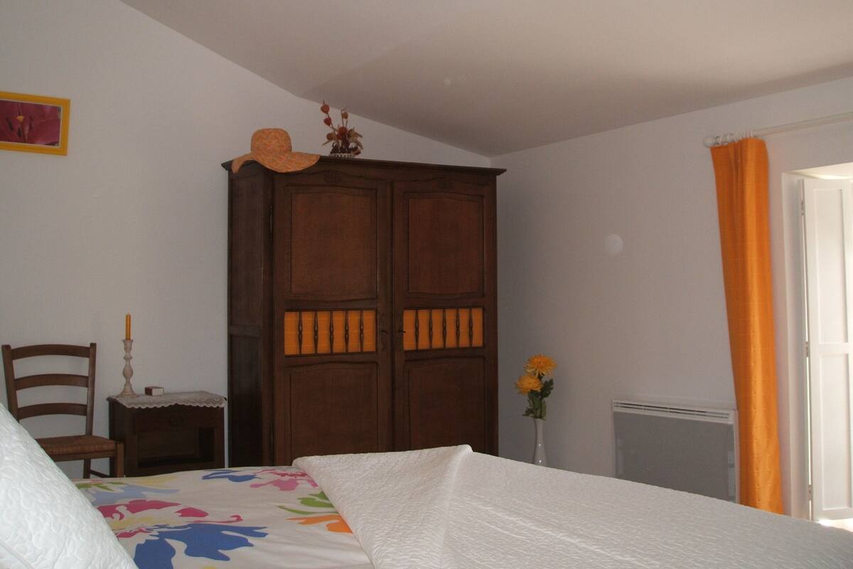 A bedroom with a bed, a wooden wardrobe, a chair, a nightstand, a vase with flowers, and a window with curtains.