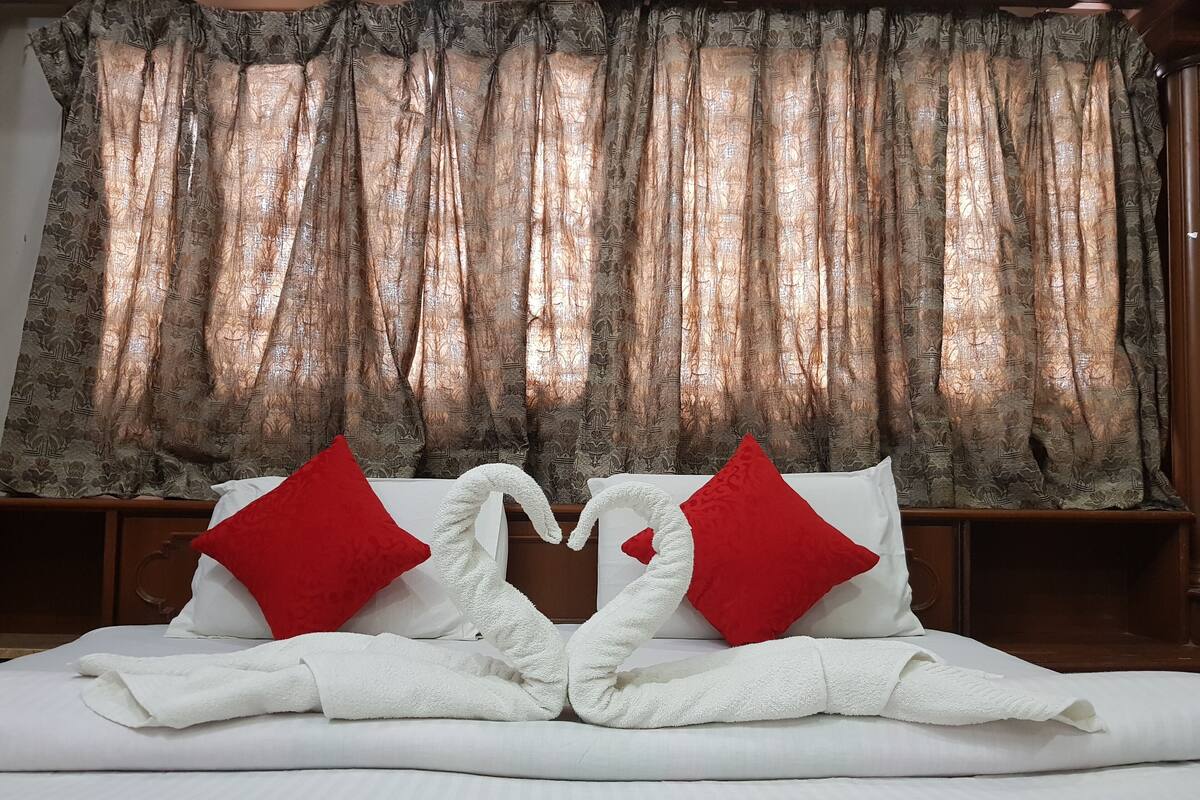 Un lit bien fait, avec du linge de lit blanc et des coussins rouges, une grande fenêtre ornée de rideaux à motifs et une tête de lit en bois.