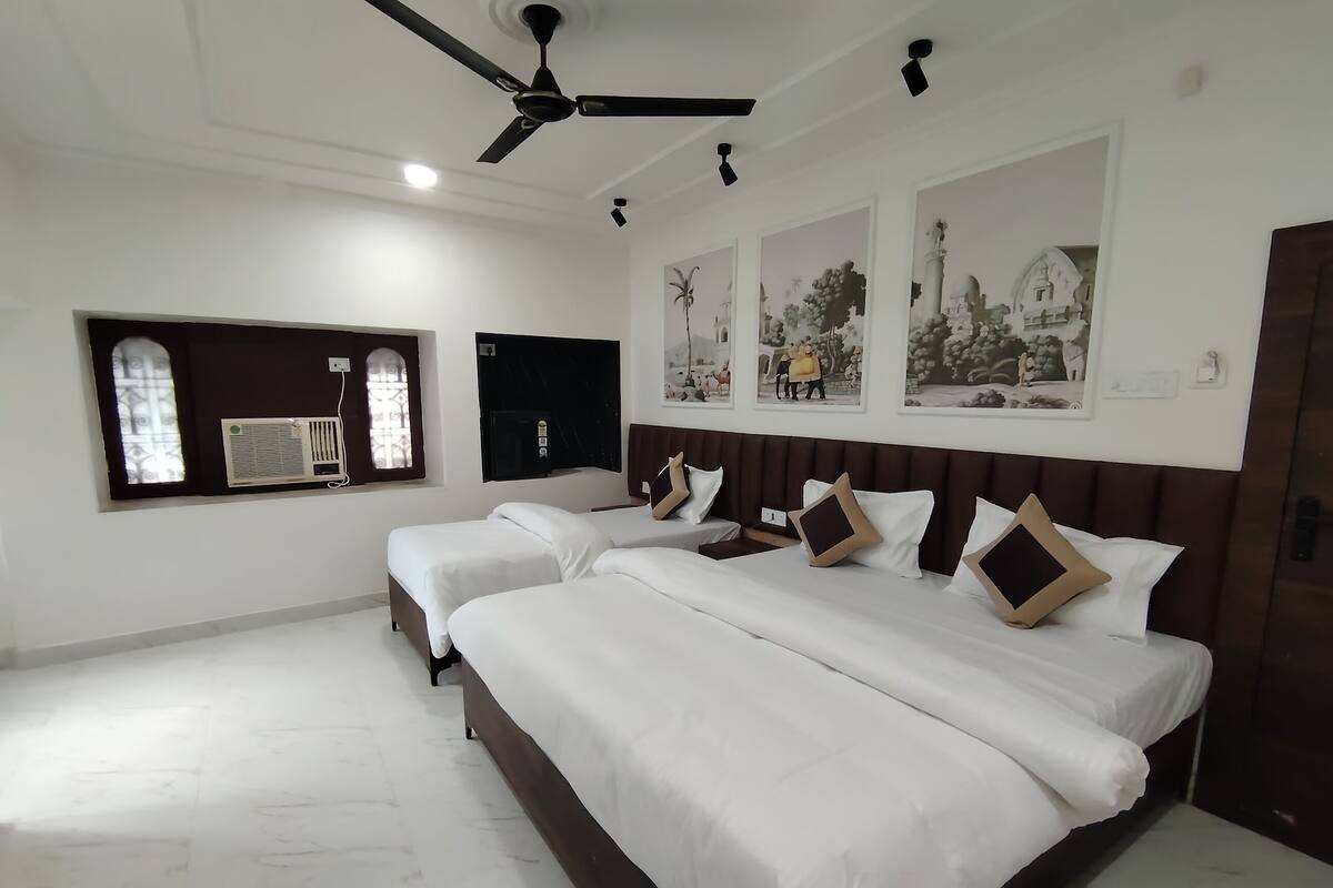 Une chambre d’hôtel avec deux lits, un ventilateur de plafond, une fenêtre avec un climatiseur et des œuvres d’art accrochées au mur.