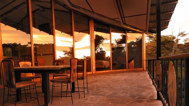 Eine Terrasse mit Steinböden, einem Tisch und Stühlen, großen Glasüren und einem hölzernen Geländer.
