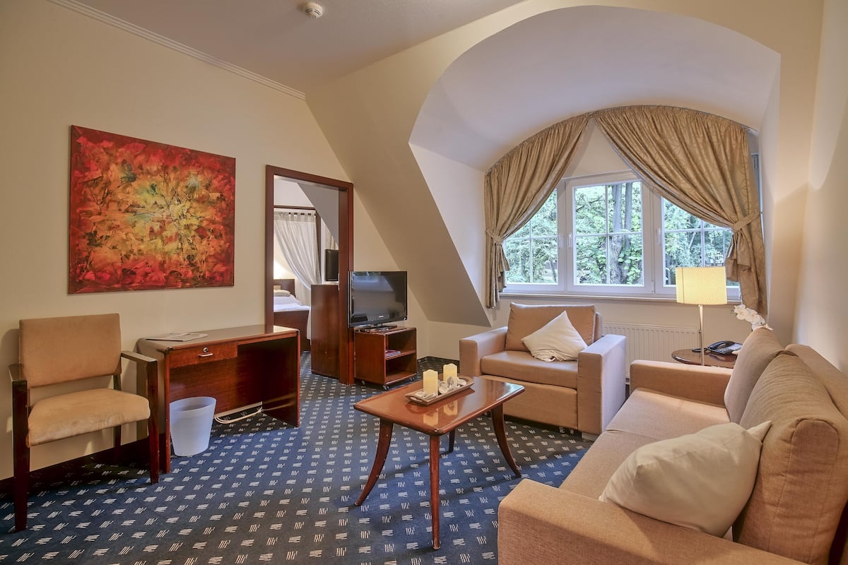 Quarto de hotel com sofá, poltrona, escrivaninha, televisão e uma janela ampla com cortinas.
